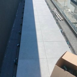 PRO - Gres - Układanie jasnoszarych płytek wielkoformatowych na długim, wąskim tarasie lub balkonie. Widoczne klipsy poziomujące, otwarty karton, szklana balustrada i ciemna ściana. Słońce tworzy wyraźny cień.