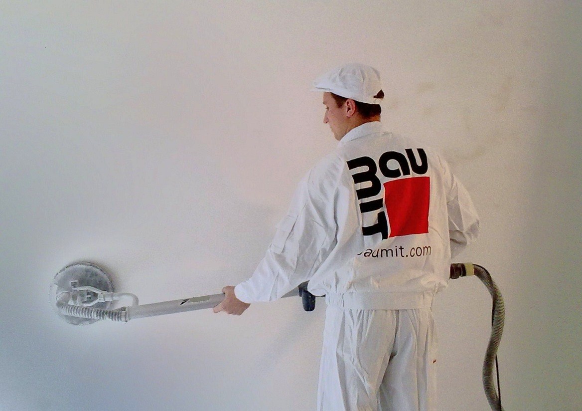 Pracownik w białym kombinezonie i czapce szlifuje ścianę za pomocą szlifierki z odkurzaczem; na plecach kombinezonu widoczne logo firmy Baumit.