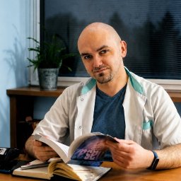 Gabinet Masażu REHKOS Krzysztof Szpakowski - Łysy mężczyzna w białym kitlu i niebieskiej koszulce, z brodą, siedzi przy drewnianym biurku, czytając otwartą książkę. W tle okno z żaluzjami i roślina. Uśmiecha się.