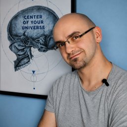 Gabinet Masażu REHKOS Krzysztof Szpakowski - Uśmiechnięty mężczyzna w okularach i szarej koszulce, z mikrofonem, na tle niebieskiej ściany i plakatu z czaszką oraz napisem 'CENTER OF YOUR UNIVERSE'.