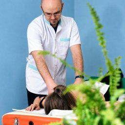 Gabinet Masażu REHKOS Krzysztof Szpakowski - Mężczyzna w białym uniformie z niebieskimi detalami, w okularach, wykonuje manualną pracę na karku osoby leżącej na pomarańczowo-drewnianym stole. Rozmyta zieleń na pierwszym planie.
