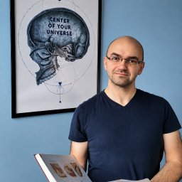 Gabinet Masażu REHKOS Krzysztof Szpakowski - Uśmiechnięty mężczyzna w okularach, trzymający otwartą książkę z anatomią, na tle niebieskiej ściany z plakatem czaszki 'CENTER OF YOUR UNIVERSE'.