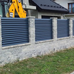 Automatyka do bram i ogrodzenia - Roman Sulejewski - Nowoczesne ogrodzenie posesji z szarych, fakturowanych bloczków betonowych i ciemnych paneli żaluzjowych, z bramą i domem w tle, częściowo widoczna koparka.