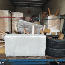 PrzeprowadzkiNowyRozdział - Załadowane wnętrze furgonetki: meble, pudła i opony owinięte folią ochronną, zabezpieczone pasami. W tle czerwony znak 'STOP'. Widoczna niebieska platforma załadunkowa.