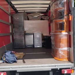 PrzeprowadzkiNowyRozdział - Wnętrze busa transportowego z załadowanymi meblami i sprzętem AGD, w tym lodówką, pralką, materacem i charakterystyczną drewnianą witryną, wszystko szczelnie owinięte folią stretch.