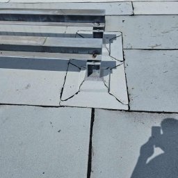 DachBudFranczak - Płaski dach pokryty jasnoszarą papą, z widocznymi pęknięciami i zużyciem wokół metalowej konstrukcji wsporczej, prawdopodobnie pod panele fotowoltaiczne.