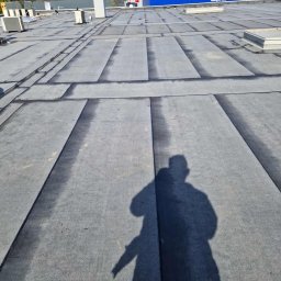 DachBudFranczak - Płaski dach pokryty ciemnoszarą membraną z widocznymi, uszczelnionymi łączeniami. Na dachu urządzenia wentylacyjne, w tle bezlistne drzewa i niebieski budynek. Widoczny cień osoby.