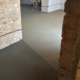 DachBudFranczak - Świeżo wylana, idealnie gładka posadzka betonowa w remontowanym wnętrzu, z widoczną zacieraczką, ścianami z płyt OSB i karton-gipsu, ujęcie zza ceglanej ściany.