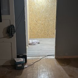 DachBudFranczak - Otwór drzwiowy w trakcie remontu, prowadzący do pomieszczenia ze ścianami z płyt OSB i wiszącą żarówką, na pierwszym planie stare deski podłogowe.