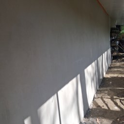 Janusz Pawlak - Długa, jasnoszara ściana zewnętrzna z widocznymi cieniami, prawdopodobnie po tynkowaniu. Na styku z sufitem pomarańczowa taśma, a na ziemi gruz budowlany.