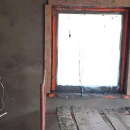 Janusz Pawlak - Świeżo otynkowana ściana z otworem okiennym zabezpieczonym folią i drewnianą ościeżnicą, obok puszki elektryczne i fragment rusztowania budowlanego.