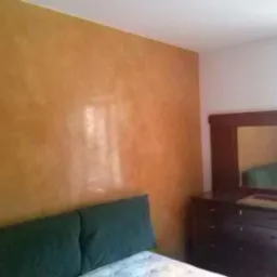 Ściana z efektem dekoracyjnym w odcieniach brązu i beżu w sypialni, widoczna komoda z lustrem i fragment łóżka, ukazująca wykończenie wnętrza.