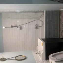 Nowoczesna łazienka po remoncie z białą armaturą, szklaną kabiną prysznicową, szarą glazurą w geometryczne wzory i dekoracyjną lampką na blacie.