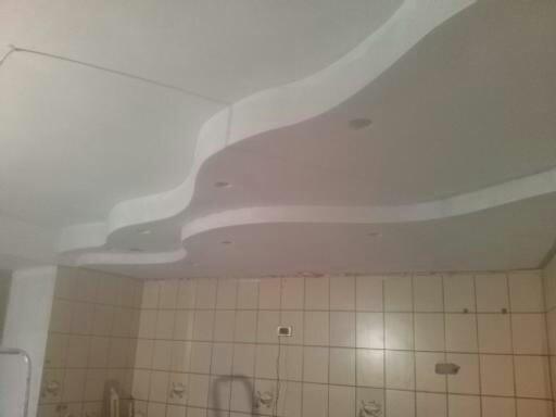 Biały sufit z dekoracyjnymi, falistymi gładziami i punktowym oświetleniem w pomieszczeniu z wyłożonymi kafelkami ścianami. Widok na wykończone wnętrze.