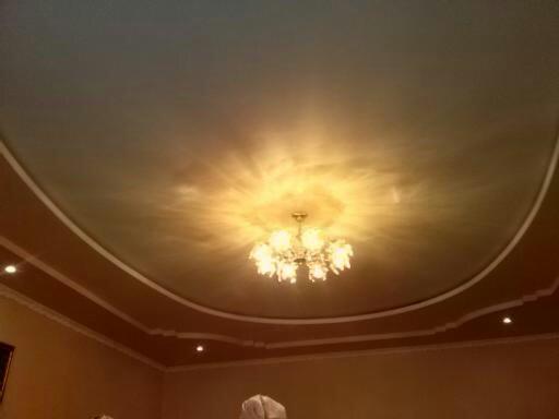 Stylowy sufit z dekoracyjnym oświetleniem i listwami ozdobnymi, centralnie umieszczony żyrandol rozświetla pomieszczenie. Widoczne detale wykończenia wnętrza.