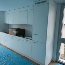 Knop - Minimalistyczna biała zabudowa kuchenna z wbudowanym piekarnikiem i miejscem na zlew, w trakcie montażu. Na blacie karton BORA, podłoga zabezpieczona niebieską folią. Widoczne dwa okna.