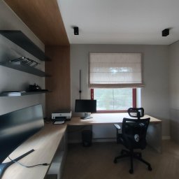 Knop - Nowoczesne biuro z jasnym, drewnianym biurkiem w kształcie litery L, ergonomicznym fotelem i dwoma monitorami. Ciemne półki ścienne i roleta rzymska nad oknem z czerwoną ramą.