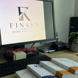 Biuro rachunkowe Finkey - Biurko z monitorem wyświetlającym logo 'FINKEY BIURO RACHUNKOWE', obok stos książek prawniczych i podatkowych, klawiatura, kalkulator oraz zielona roślina.