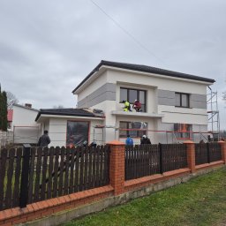 Piotr Wlazło - Nowoczesny dom z beżową elewacją i szarymi pasami, gdzie pracownicy na rusztowaniach wykańczają detale przy oknach. Widoczne zabezpieczone okna i drewniane ogrodzenie z ceglanymi słupkami.