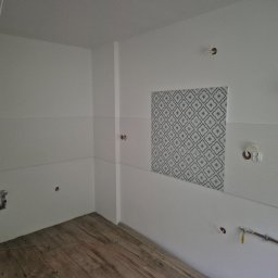 Piotr Wlazło - Wnętrze remontowanego pomieszczenia z białymi ścianami, częściowo pokrytymi wzorzystymi płytkami geometrycznymi i białymi ryflowanymi. Widoczne instalacje elektryczne, rury hydrauliczne i drewniana...