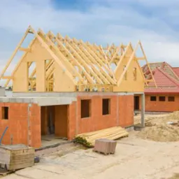 Budowa domu w stanie surowym zamkniętym: ściany z czerwonej cegły, drewniana konstrukcja dachu w trakcie montażu, widoczne deski i bloczki budowlane na placu budowy.