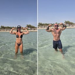 FIT A&M - Kobieta w bikini i czapce 'NY' oraz mężczyzna w kąpielówkach, oboje z tatuażami, prężą mięśnie, stojąc w płytkiej, turkusowej wodzie przy piaszczystej plaży z palmami.
