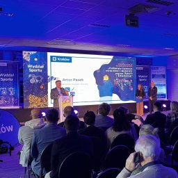 Gremium - Realizacja Techniczna i Studio Nagrań - Scena konferencyjna z prelegentem Arturem Paskiem przy podium, oświetlona niebieskim światłem. Na dużym ekranie LED widnieje tytuł 'Konferencja SZKOLENIOWA: Profilaktyka Uzależnień...' oraz logo...