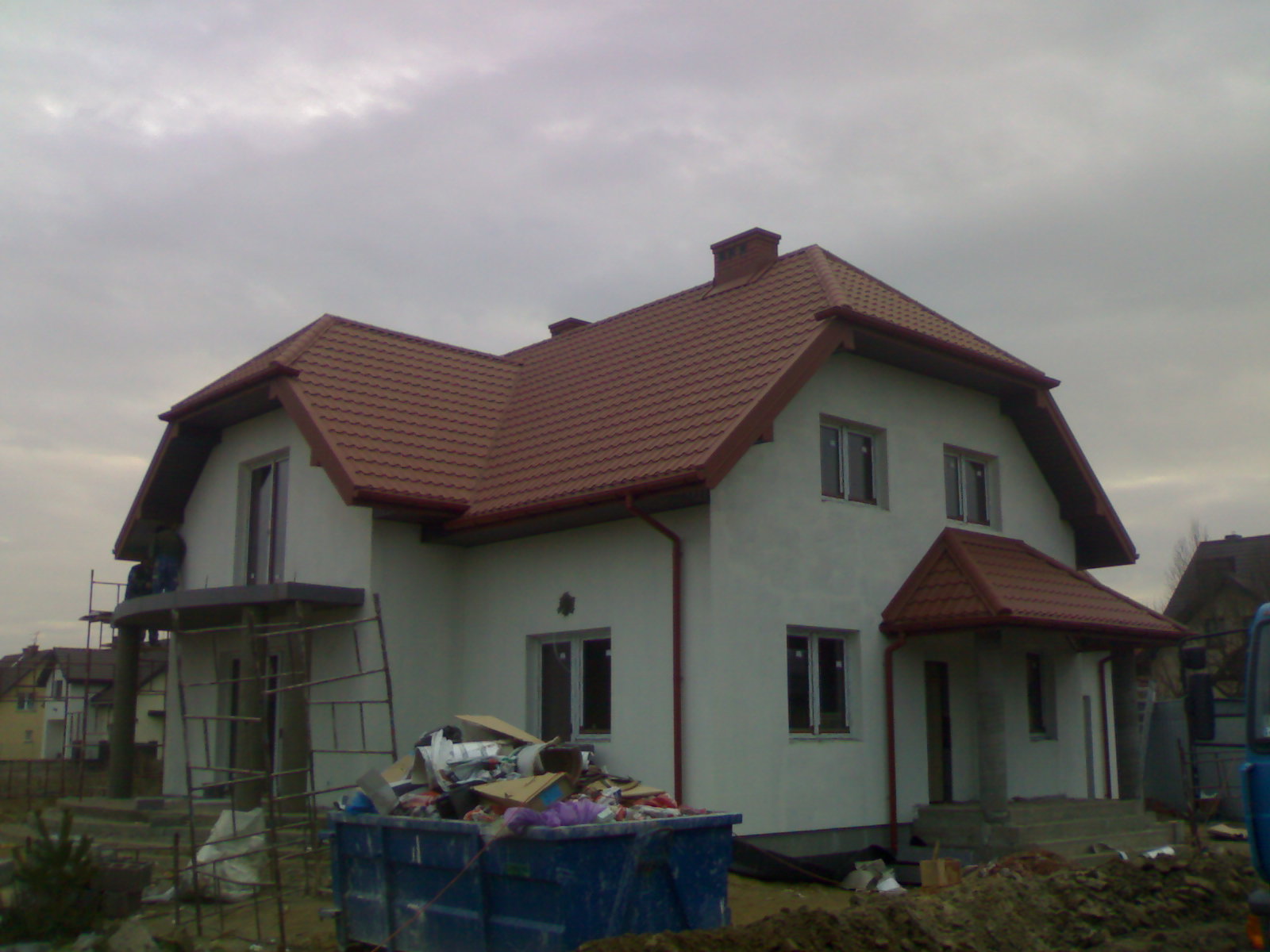 Nowy dom jednorodzinny z dachem pokrytym brązową blachodachówką, widoczny kontener ze śmieciami budowlanymi i rusztowanie przy balkonie.