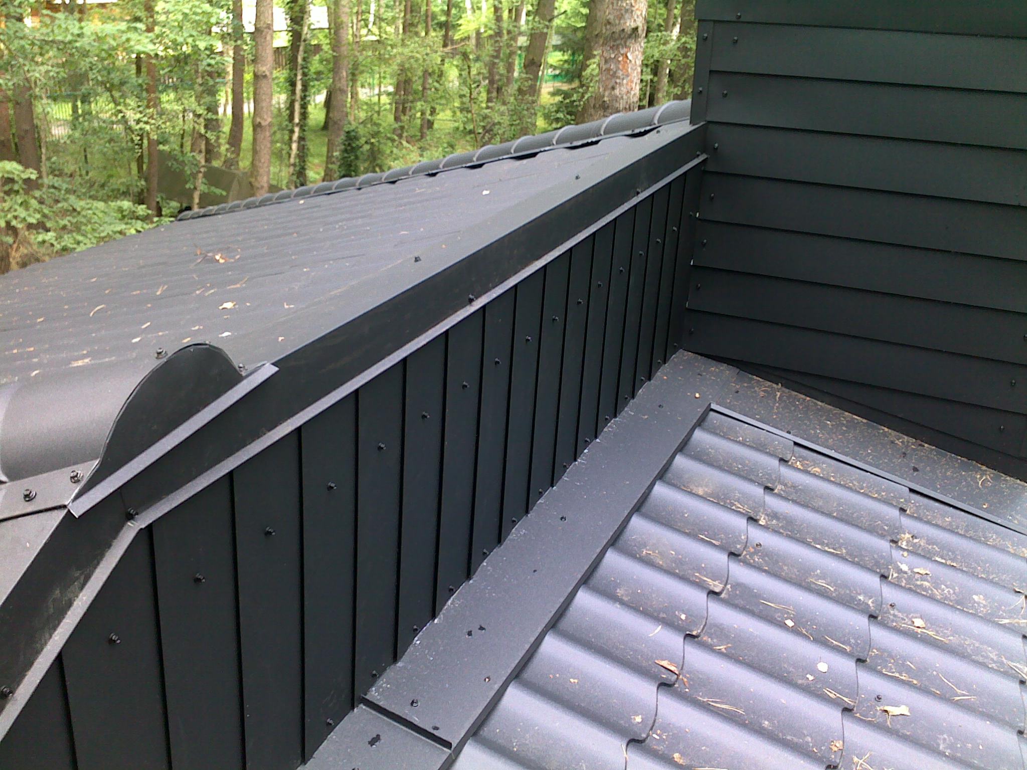 Fragment dachu pokrytego ciemną blachodachówką z widocznymi elementami obróbek blacharskich i drewnianą obudową komina na tle drzew.