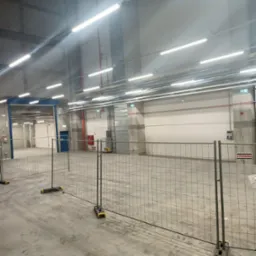 Wnętrze hali magazynowej z widocznymi lampami jarzeniowymi pod sufitem, instalacją przeciwpożarową w kolorze czerwonym, szarą podłogą betonową, zabezpieczoną barierkami ochronnymi w kolorze szarym...