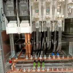 Wnętrze szafy elektrycznej z grubymi kablami oznaczonymi L1, L2, L3 podłączonymi do miedzianych szyn zbiorczych, widoczne bezpieczniki i inne elementy instalacji.