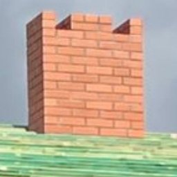 Remont i naprawa dachów i elewacji - Solidny ceglany komin z dekoracyjnym, schodkowym zwieńczeniem, wznoszący się ponad zielonymi łatami dachu, na tle pochmurnego nieba.