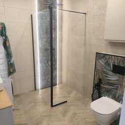 REMO - Nowoczesna łazienka z kabiną walk-in z czarną ramą, liniowym oświetleniem LED i płytkami z tropikalnym wzorem. Podłoga z płytek drewnopodobnych w jodełkę.