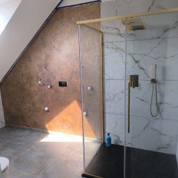 REMO - Nowoczesna łazienka z kabiną prysznicową ze złotymi okuciami. Ściana z efektem miedzi i widocznymi instalacjami, obok białe płytki imitujące marmur. Szare płytki podłogowe.