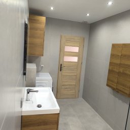REMO - Nowoczesna łazienka z umywalką na szafce w kolorze jasnego drewna, pralką w zabudowie, drzwiami z przeszkleniami i szarymi płytkami na ścianach i podłodze.