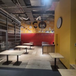 REMO - Nowoczesne wnętrze lokalu gastronomicznego z industrialnym sufitem, żółtymi ścianami z grafikami 'CHAPS GA-ROPUB Poznań' i czerwoną, falistą ścianą. Jasne stoły, lada z czarno-białymi płytkami...