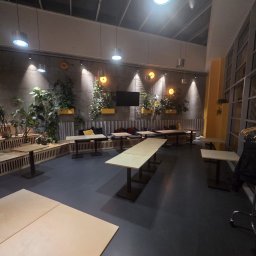 REMO - Nowoczesne wnętrze z betonową ścianą, podwieszanymi żółtymi donicami z bujnymi roślinami, długimi drewnianymi stołami i ciemną podłogą. Industrialne oświetlenie punktowe i zabudowa z drewna.