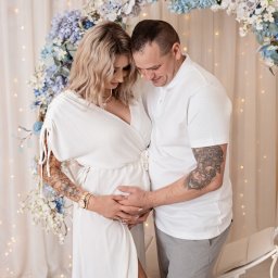 Studio Fotoszpulka - Przyszli rodzice w białych strojach, czule obejmujący ciążowy brzuszek, na tle jasnych zasłon i okrągłej dekoracji z niebiesko-białych kwiatów.