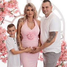 Studio Fotoszpulka - Rodzina pozuje w studio: kobieta w różowej sukience z różą na brzuchu i tatuażami, mężczyzna z tatuażem i chłopiec, z dłońmi na jej brzuchu, na tle białej ramy i różowych kwiatów.