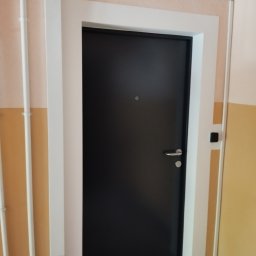 Bodeko - Ciemne drzwi wejściowe z srebrną klamką i wizjerem, osadzone w białej ościeżnicy, na tle dwukolorowej ściany (jasny róż i żółty). Widoczne białe rurki instalacyjne po lewej.