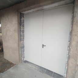 Bodeko - Nowo zamontowane, jasne dwuskrzydłowe drzwi z czarną klamką, osadzone w surowym otworze z betonu i cegły, z betonową posadzką.