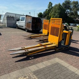 WIDLAKI69 - Niemieckie Centrum Szkoleniowe - Żółty elektryczny wózek paletowy z długimi widłami, zaparkowany na brukowanym placu. W tle biały van dostawczy z otwartymi drzwiami i biały SUV w słoneczny dzień.