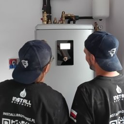 InstallBrothers - Dwóch instalatorów w firmowych koszulkach, tyłem do kamery, analizuje nowoczesny system grzewczy z cyfrowym panelem sterowania i mosiężnymi przyłączami.