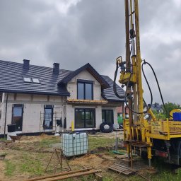 InstallBrothers - Duża żółta wiertnica wykonująca odwierty pod systemy grzewcze, z rurami na ziemi, obok budowanego domu z czarnym dachem i oknami.