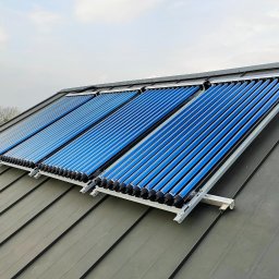 InstallBrothers - Trzy rzędy niebieskich kolektorów słonecznych próżniowych PROMAG, zamontowanych na ciemnoszarym, skośnym dachu z blachodachówki, pod pochmurnym niebem.