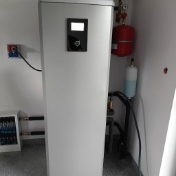 InstallBrothers - Nowoczesne urządzenie grzewcze Thermia z panelem sterowania, mosiężnymi rurami, czerwonym naczyniem wzbiorczym i niebieskim płynem, na tle białej ściany i szarej podłogi.
