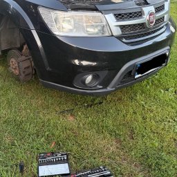 PB Mobilny Mechanik Szczecin i Okolice - Czarny Fiat Freemont z otwartą maską i zdjętym przednim lewym kołem, ukazujący tarczę hamulcową. Na trawie leżą otwarte zestawy kluczy nasadowych, przygotowane do serwisu.