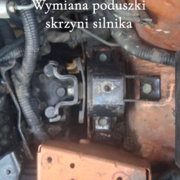 PB Mobilny Mechanik Szczecin i Okolice - Wymiana poduszki skrzyni silnika, zbliżenie na metalowy wspornik z gumowym elementem, otoczony przewodami i rurkami w komorze silnika.
