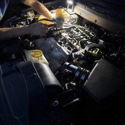 PB Mobilny Mechanik Szczecin i Okolice - Ręce osoby pracującej przy silniku samochodu, oświetlone punktowym światłem roboczym, z widocznym zbiornikiem płynu chłodniczego i elementami układu napędowego.