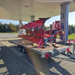 Skowroński Usługi Mini Koparką - Czerwony pług rolniczy z kilkoma lemieszami, solidnie zabezpieczony różnokolorowymi pasami transportowymi na dwuosiowej lawecie, stoi pod zadaszeniem stacji paliw z numerem 3 w Tomaszowie Lubelskim.
